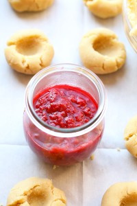 A jar of raspberry jam beside Raspberry Thumbprint Cookies on a baking sheet.