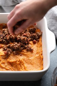 A person preparing a healthy sweet potato casserole by scooping mashed sweet potatoes into a dish.