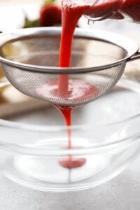 Blended strawberries being poured into a sieve over a glass bowl.