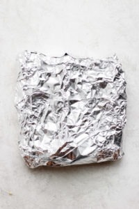 Grilled corn wrapped in aluminum foil on a white surface.
