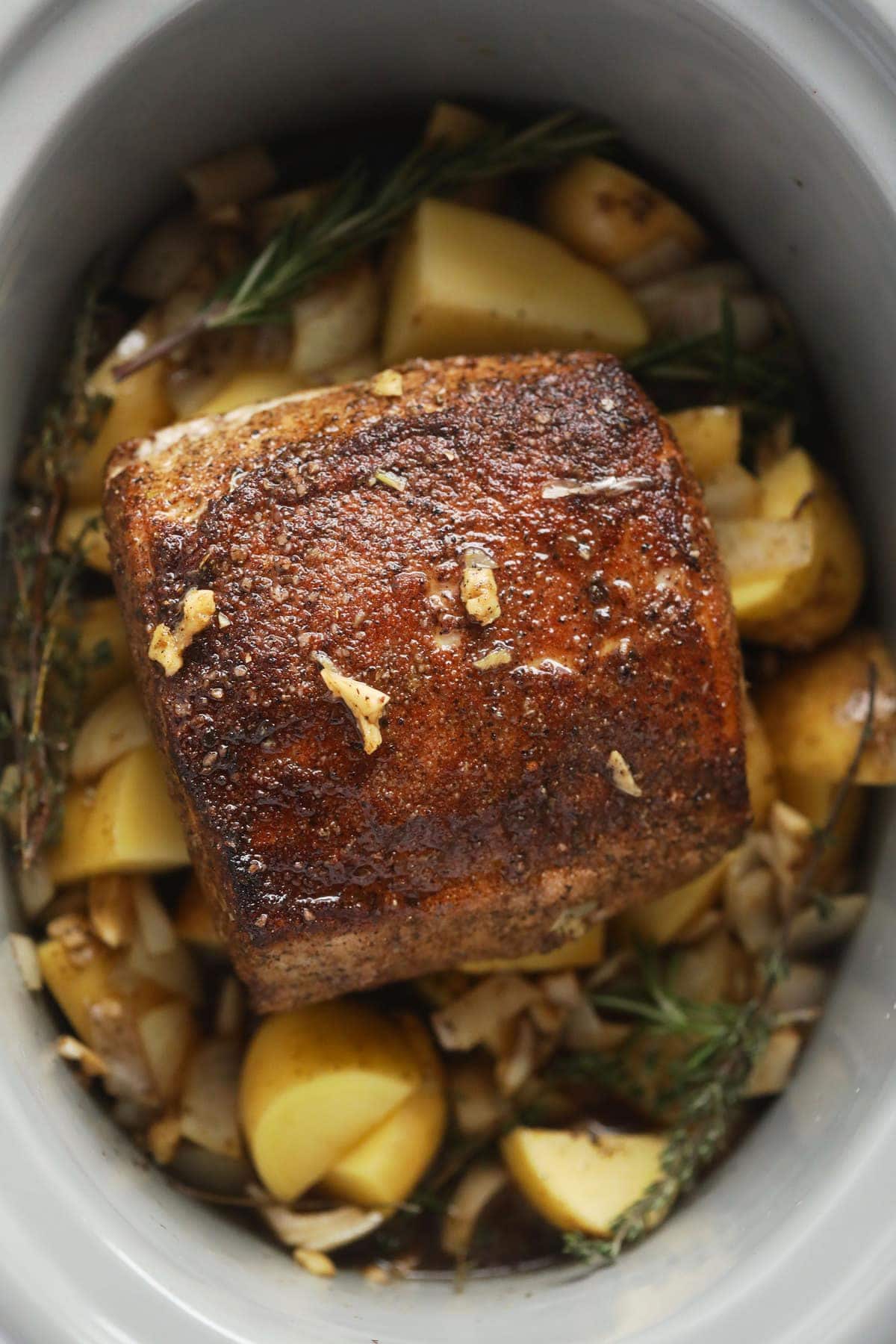 slow cooker pork loin roast on a bed of potatoes and herbs in the slow cooker.