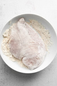 A raw chicken breast covered in flour on a white plate on a white surface.