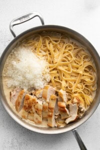 A stainless steel pan contains cooked fettuccine pasta, sliced grilled chicken breast, and grated Parmesan cheese, with a creamy sauce.