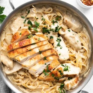 A pan filled with creamy fettuccine pasta topped with slices of grilled chicken and garnished with parsley. A small bowl of red pepper flakes is seen in the background.