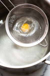 An egg is being fried in a metal strainer.