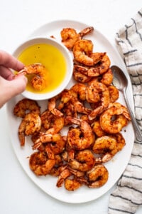 A plate of grilled shrimp with a hand holding a bowl of olive oil.