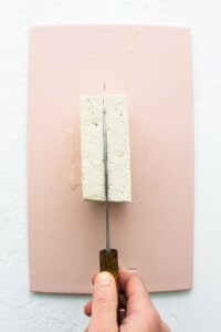 A person cutting a piece of tofu on a pink surface.