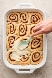 A person is dipping a spatula into a dish of cinnamon rolls.