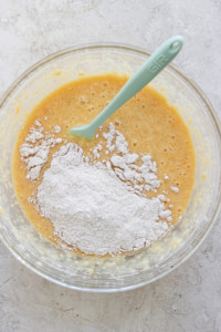 A glass bowl containing a mixture of flour and a liquid batter with a green silicone spatula.