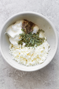 A bowl of crumbled feta cheese mixed with seasonings and yogurt on a textured surface.