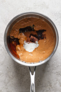 A stainless steel saucepan containing ingredients for a dish, including a reddish-brown sauce, visible spices, and a dollop of white substance, possibly coconut milk or cream, in the process of.