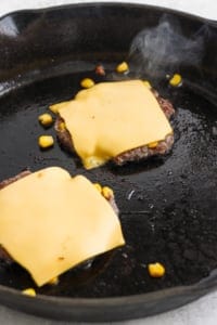Two beef patties with melted cheese slices on top cooking in a skillet.
