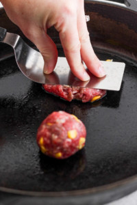A person flattening a ball of ground meat on a skillet to form a patty.