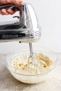A person is using a silver cuisinart hand mixer to blend batter in a clear glass bowl on a kitchen countertop.