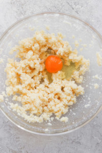 A glass bowl containing crumbled dough and a whole raw egg on a textured surface.