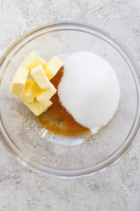 Ingredients for baking, including butter, white sugar, and brown sugar, are placed in a glass mixing bowl on a textured surface.