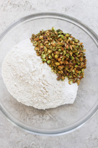 A clear glass bowl containing a pile of white flour and shelled green pistachios on a neutral surface.
