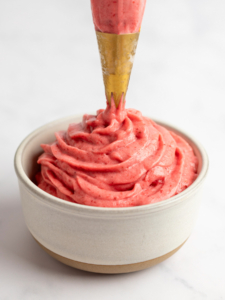 A piping bag dispensing creamy strawberry frosting into a small beige bowl against a white background.