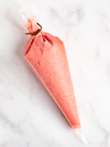 A piping bag filled with pink frosting, tied at the top, resting on a white marble surface.