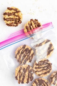 A clear plastic bag with a pink zip seal contains round cookies drizzled with chocolate. Two cookies are placed outside the bag.