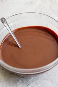 A glass bowl containing smooth melted chocolate with a metal spoon resting inside, set on a light-colored surface.