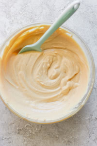 A glass bowl containing creamy peanut butter with a green spoon, photographed from overhead on a textured white surface.