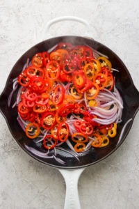 Sliced red and yellow bell peppers with red onions in a white le creuset skillet on a textured surface.
