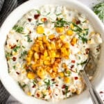 A bowl of creamy dip topped with roasted corn, chopped herbs, and red pepper flakes, with a spoon on the side.