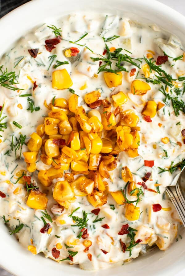 A bowl of creamy dip topped with roasted corn, chopped herbs, and red pepper flakes, with a spoon on the side.
