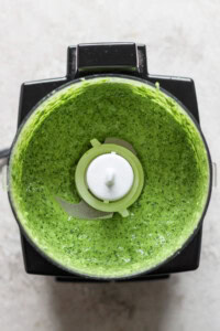 A top-down view of a blender with green sauce inside, featuring visible greens and herbs blended to a smooth consistency.