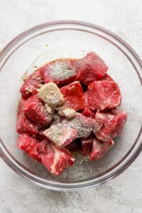 A glass bowl filled with raw beef chunks seasoned with spices on top, placed on a light-colored textured surface.