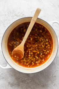 A white pot filled with a red broth containing chopped onions, stirred with a wooden spoon, placed on a gray textured surface.