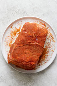 A piece of seasoned raw meat on a white plate, placed on a textured white surface.