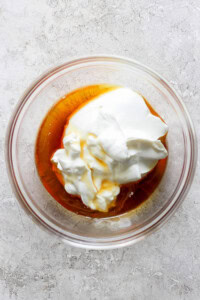 A glass bowl containing Greek yogurt and honey on a textured, light-colored surface.