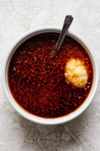 A white bowl filled with a reddish liquid and finely chopped ingredients, with a spoon and a dollop of minced garlic on top.