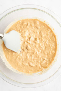 A glass bowl containing a thick, beige batter mixture with a white spatula resting on the side.