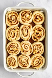 A baking dish that contains twelve unbaked cinnamon rolls arranged in rows on a light-colored surface.