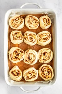 A white baking dish filled with twelve unbaked cinnamon rolls evenly spaced, ready for baking, on a white marble surface.