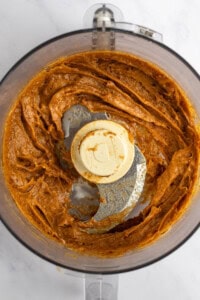 A food processor contains a blended brownish-orange mixture with a smooth consistency. The processor's blade and lid cover are visible.