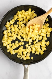 Diced potatoes cooking in a black skillet with a wooden spoon on a white marble surface.
