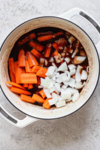 A white pot on a light surface contains chopped carrots, diced onions, minced garlic, and a dark liquid. The ingredients appear to be in the initial stages of cooking.
