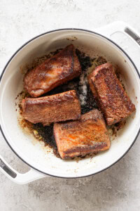 Four browned meat pieces are in a white, enameled Dutch oven on a light surface.