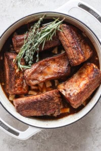 A white pot contains several pieces of cooked beef short ribs garnished with fresh rosemary and thyme.