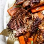 A plate of tender roasted meat with a crispy exterior, served on a bed of mashed potatoes and garnished with cooked carrots and rosemary, with two forks visible on the side.