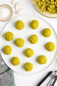A white plate with twelve round, greenish-yellow falafel balls. Nearby are a bowl of falafel mixture, a bowl of salt, three garlic cloves, a cloth, and a pair of tongs.