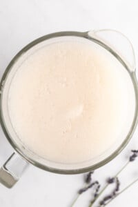 A clear glass bowl filled with a white, granular substance, viewed from above. Two sprigs of lavender are placed beside the bowl on a white surface.