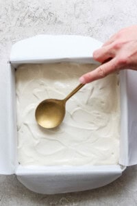 A hand holding a spoon smooths out a white batter for peanut butter banana ice box cake in a parchment-lined square baking pan on a textured countertop.