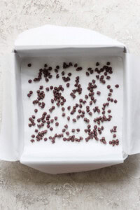 A square baking pan lined with parchment paper, scattered with small chocolate chips, hints at the indulgent layers of a peanut butter banana ice box cake, placed on a light-colored surface.
