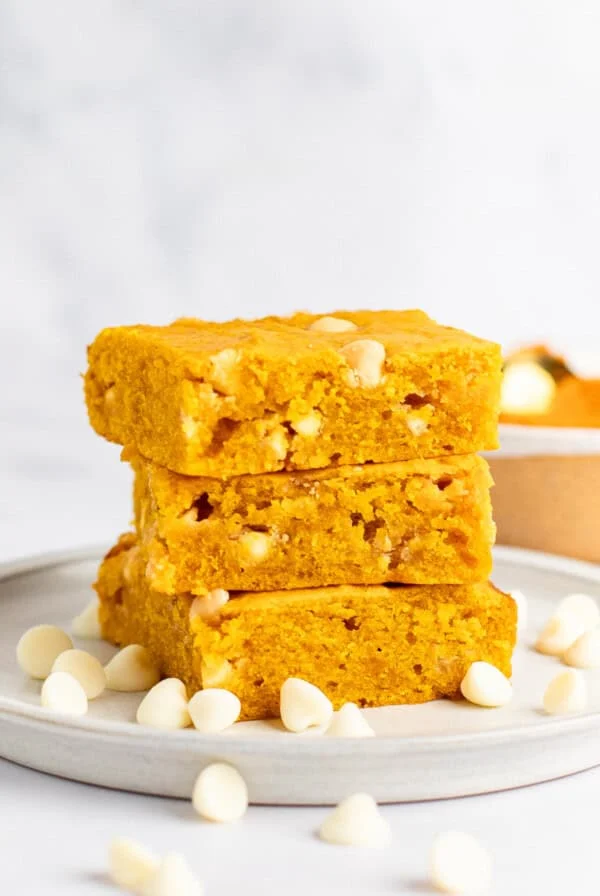 Three slices of golden yellow pumpkin blondies stacked on a plate, surrounded by white chocolate chips.