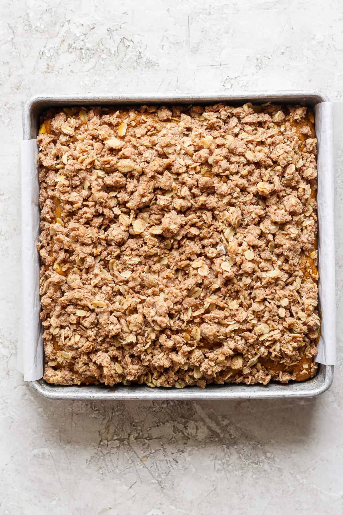 A square metal baking pan filled with a baked oatmeal fruit crumble topped with a crispy, golden-brown oat and cinnamon streusel. The pan is lined with parchment paper.